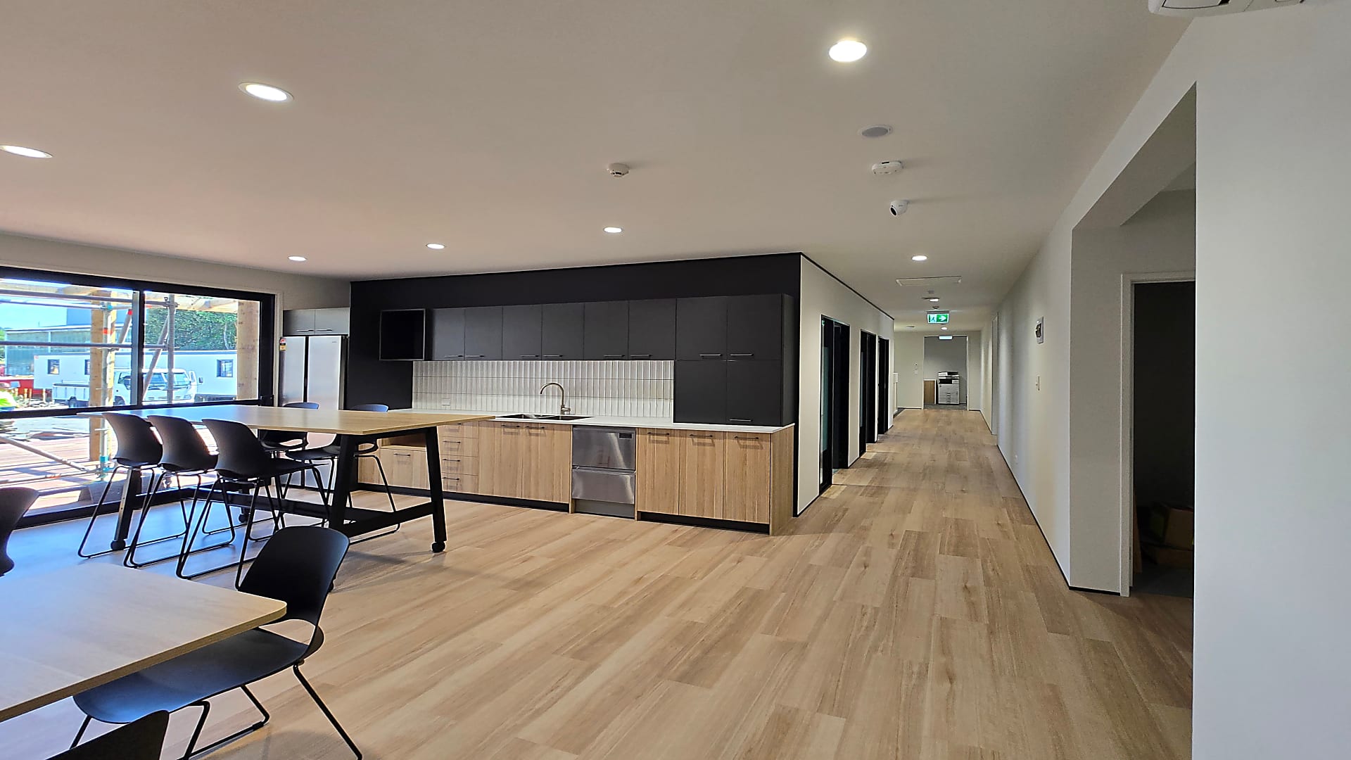 Modern office kitchenette with wooden lower cabinets, black upper cabinets, stainless steel appliances, high table with black chairs, and a long hallway with wooden flooring.