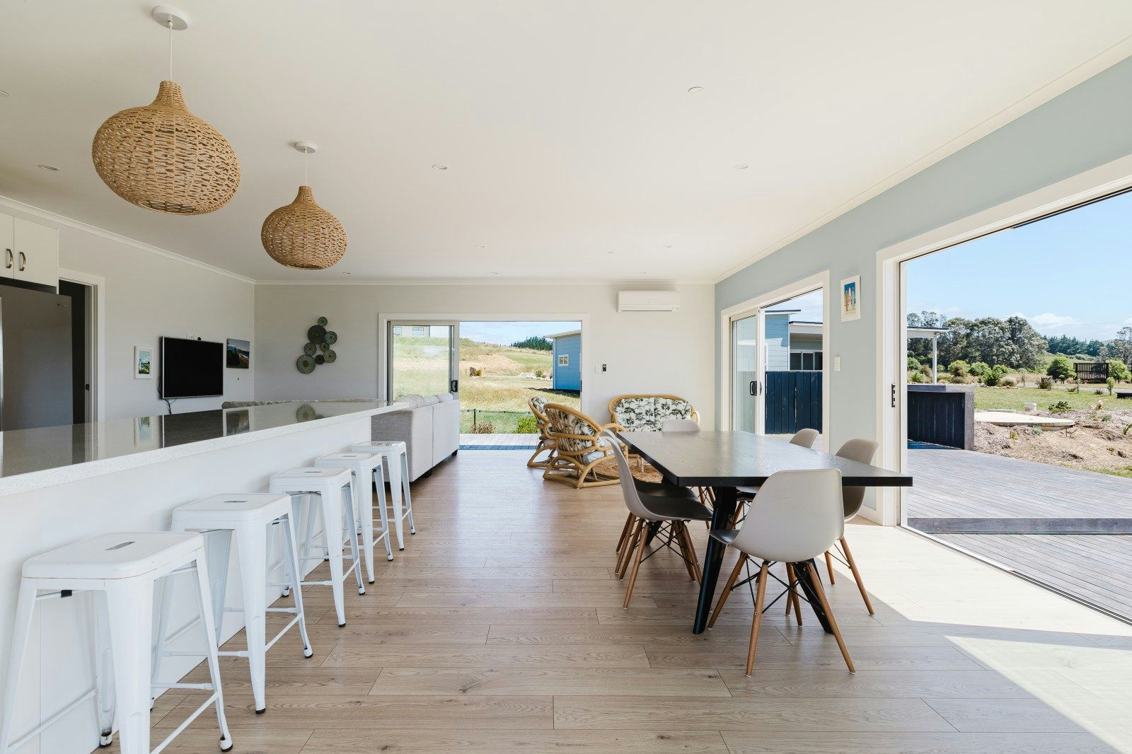 Bright open kitchen and dining area with white bar stools, black dining table, modern chairs, and large sliding doors opening to a wooden deck with greenery outside.