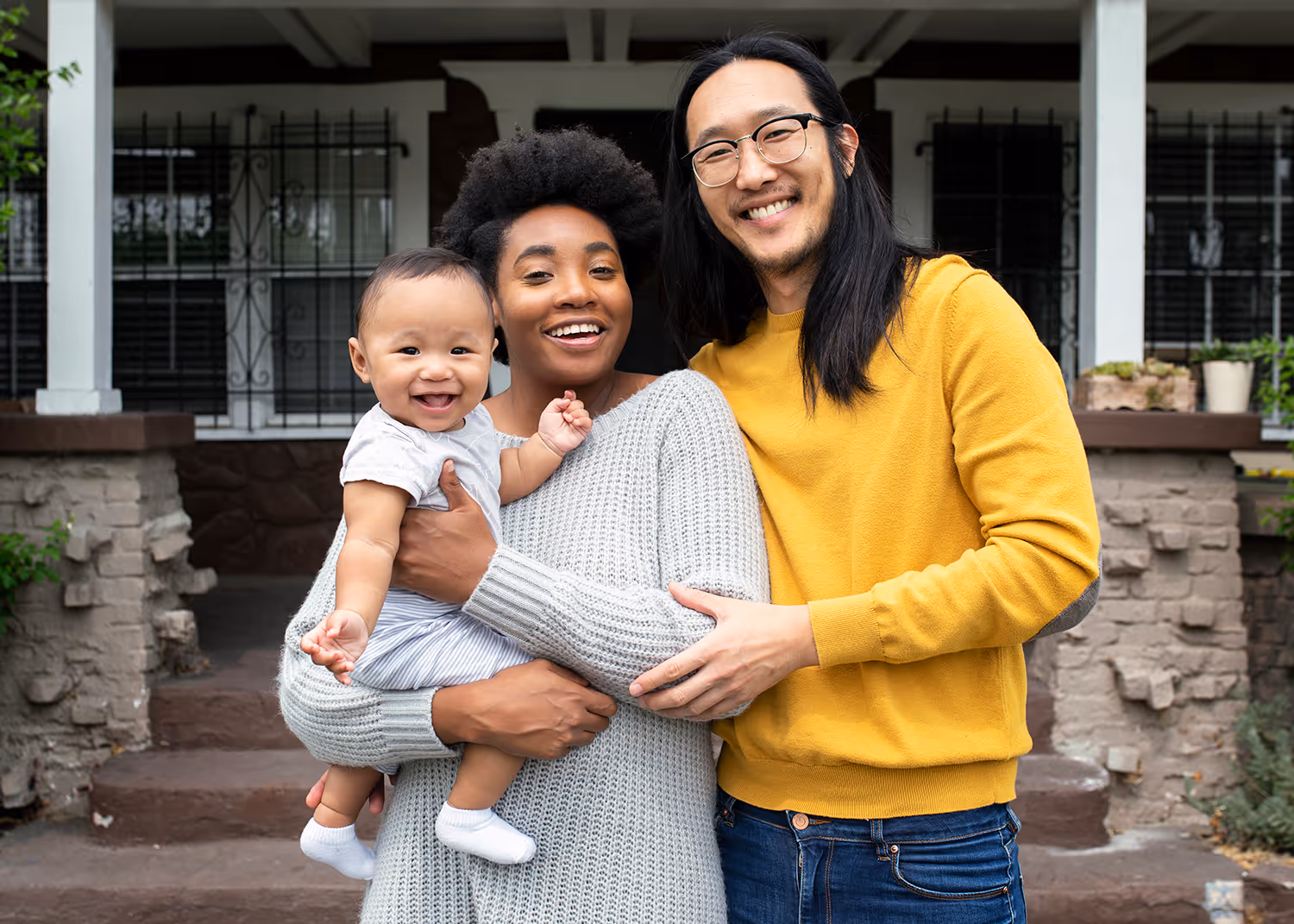 Smiling interracial couple standing on a porch with the woman holding a happy baby.
