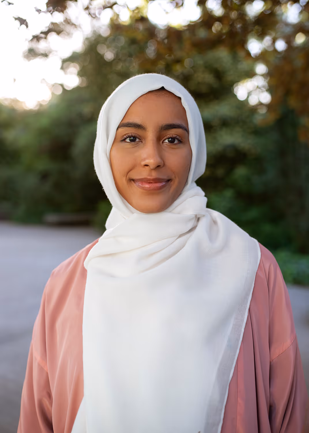 Smiling woman wearing a white hijab and peach blouse standing outdoors with greenery in the background.