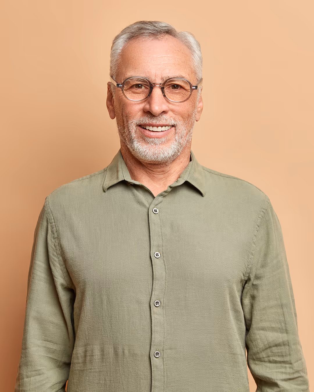 Smiling elderly man with gray hair and beard, wearing glasses and a light green buttoned shirt, against a tan background.