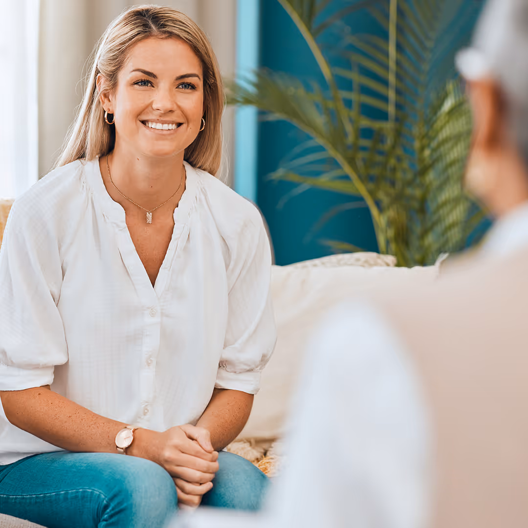 Smiling woman with blonde hair wearing a white blouse and blue jeans, sitting and engaging in conversation with a person in the foreground.