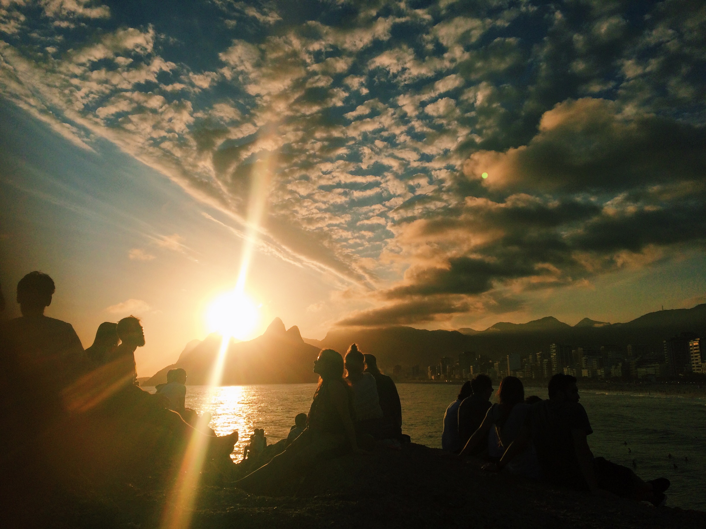 People watch the sunset behind Dois Irmãos mountain sitting on Arpoador rock