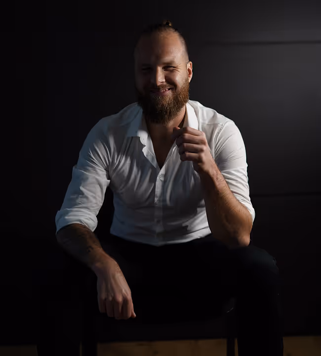 Bearded man with a bun hairstyle sitting on a chair against a dark background, wearing a white shirt and smiling.