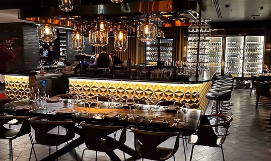 Elegant dimly lit restaurant interior with a bar featuring modern geometric lighting, bar stools, and tables set with glassware and plates.