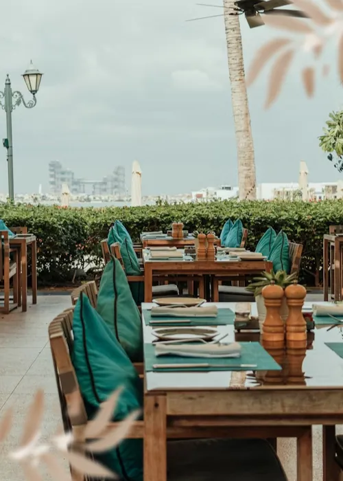 Outdoor restaurant seating area with wooden tables set with napkins, cutlery, and salt and pepper shakers, teal cushions on chairs, and a background of greenery and overcast sky.