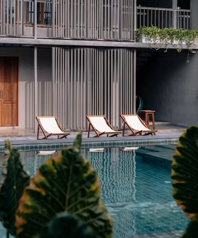 Three wooden lounge chairs with white fabric lined up beside a clear outdoor swimming pool in front of a modern building with vertical slatted walls and a balcony with green hanging plants.
