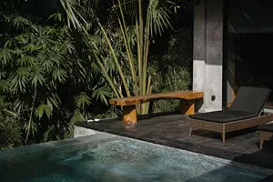Wooden bench and wicker lounge chair beside a pool with tropical plants in the background at night.