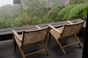 Two wooden chairs with woven backrests on a dark wooden deck overlooking lush green foliage and trees in a misty environment.