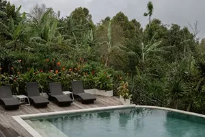 Swimming pool with several black lounge chairs on a wooden deck surrounded by lush tropical greenery under a cloudy sky.