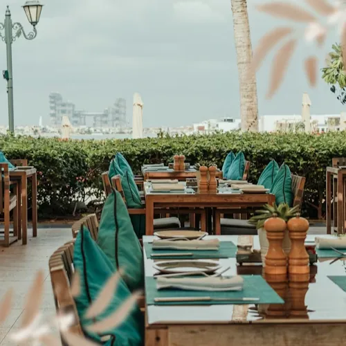 Outdoor restaurant seating area with wooden tables set with napkins, cutlery, and salt and pepper shakers, teal cushions on chairs, and a background of greenery and overcast sky.