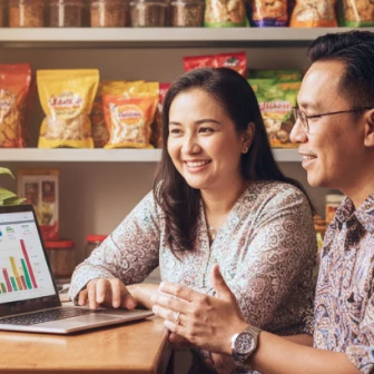 Two people smiling and discussing data shown on a laptop screen in a room with shelves stocked with packaged food products.