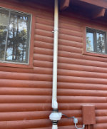 White vent pipe running vertically against a brown log cabin exterior with two windows.
