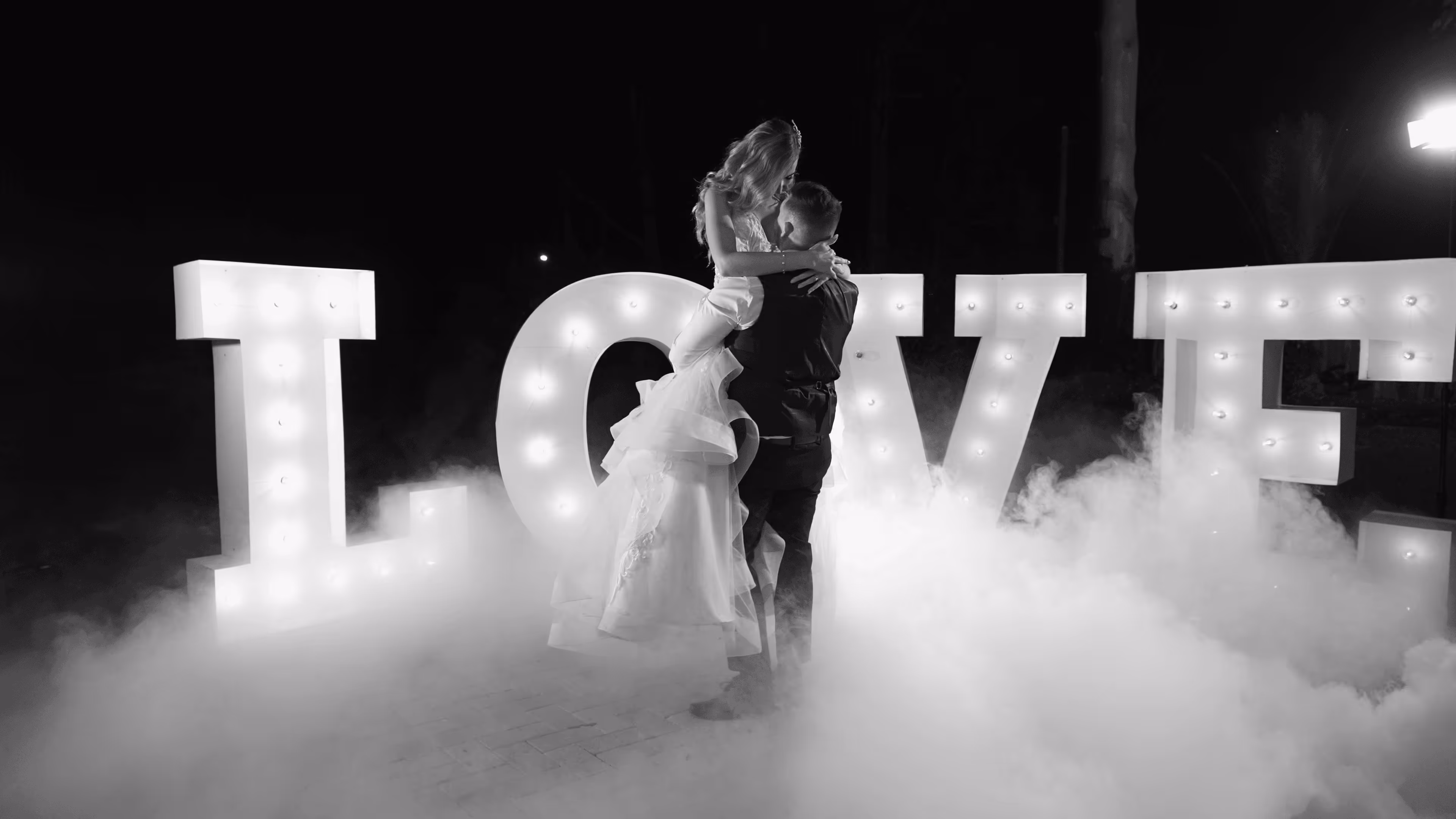 Bride and groom share a joyful lift surrounded by glowing marquee letters spelling LOVE and a swirl of soft mist.