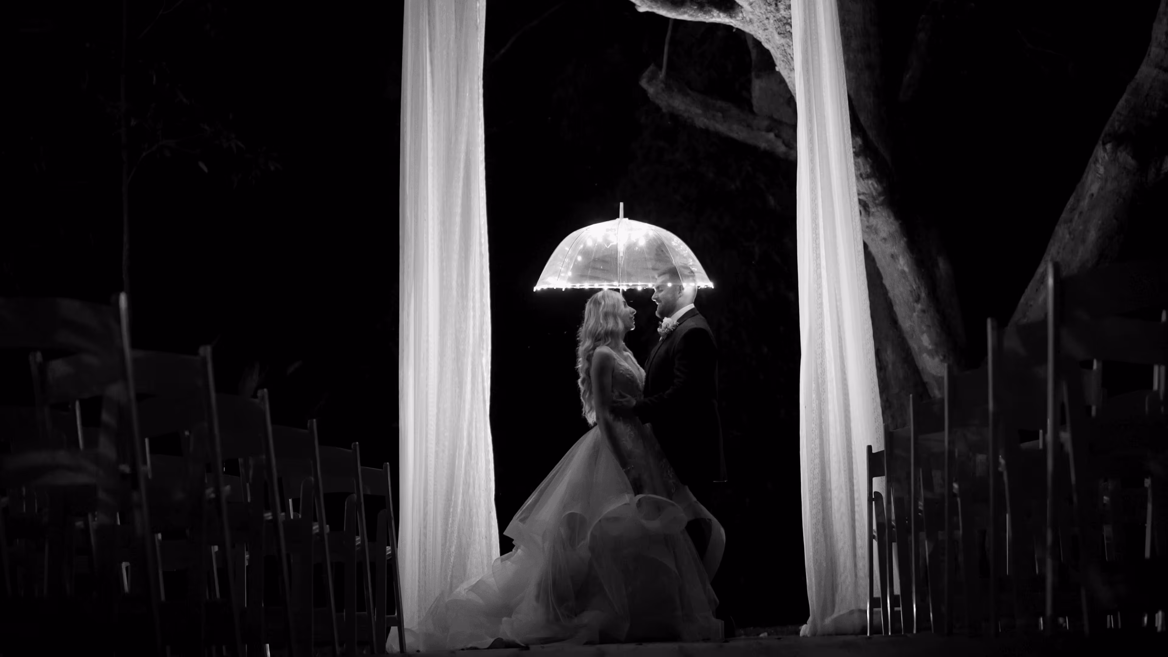 Bride and groom stand beneath flowing drapes under a clear umbrella, softly illuminated in the night during a rain-kissed ceremony.