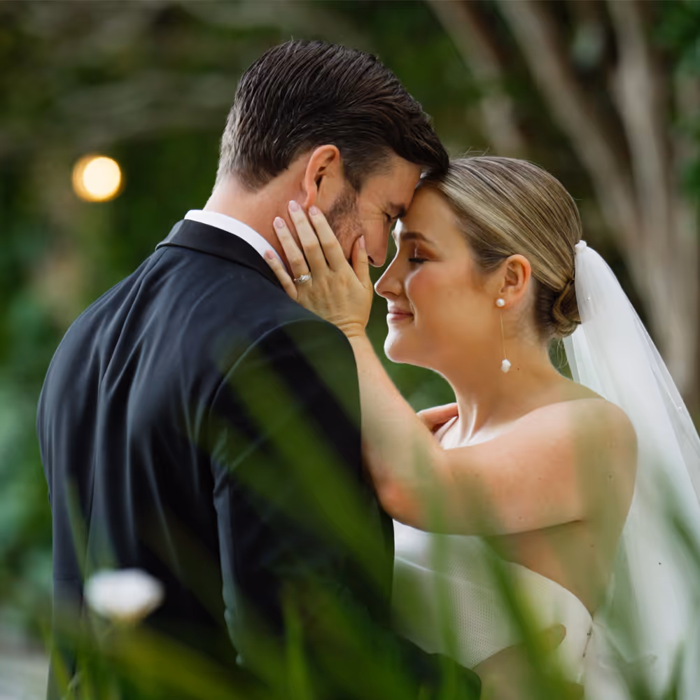 Bride and groom share a quiet forehead embrace surrounded by soft greenery, their eyes closed in a moment of calm connection.