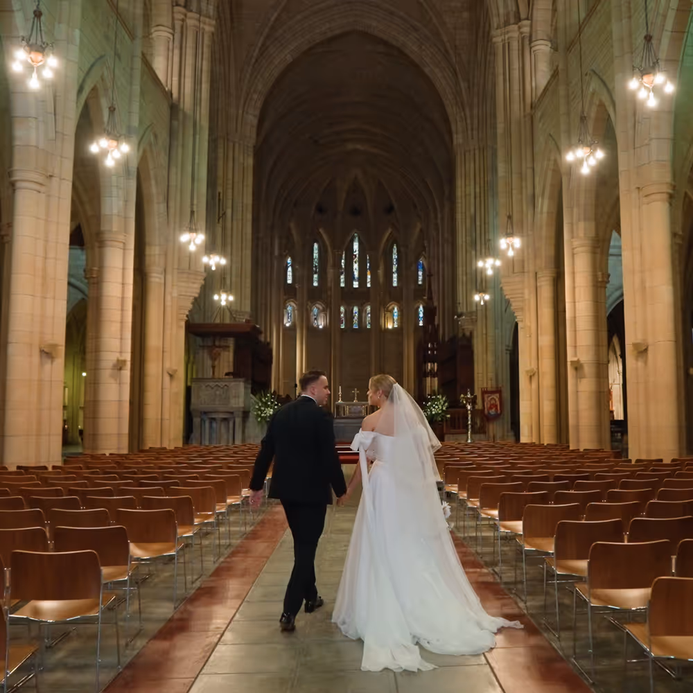 Bride and groom walk hand in hand down the aisle of a grand cathedral, framed by soaring arches and golden light.