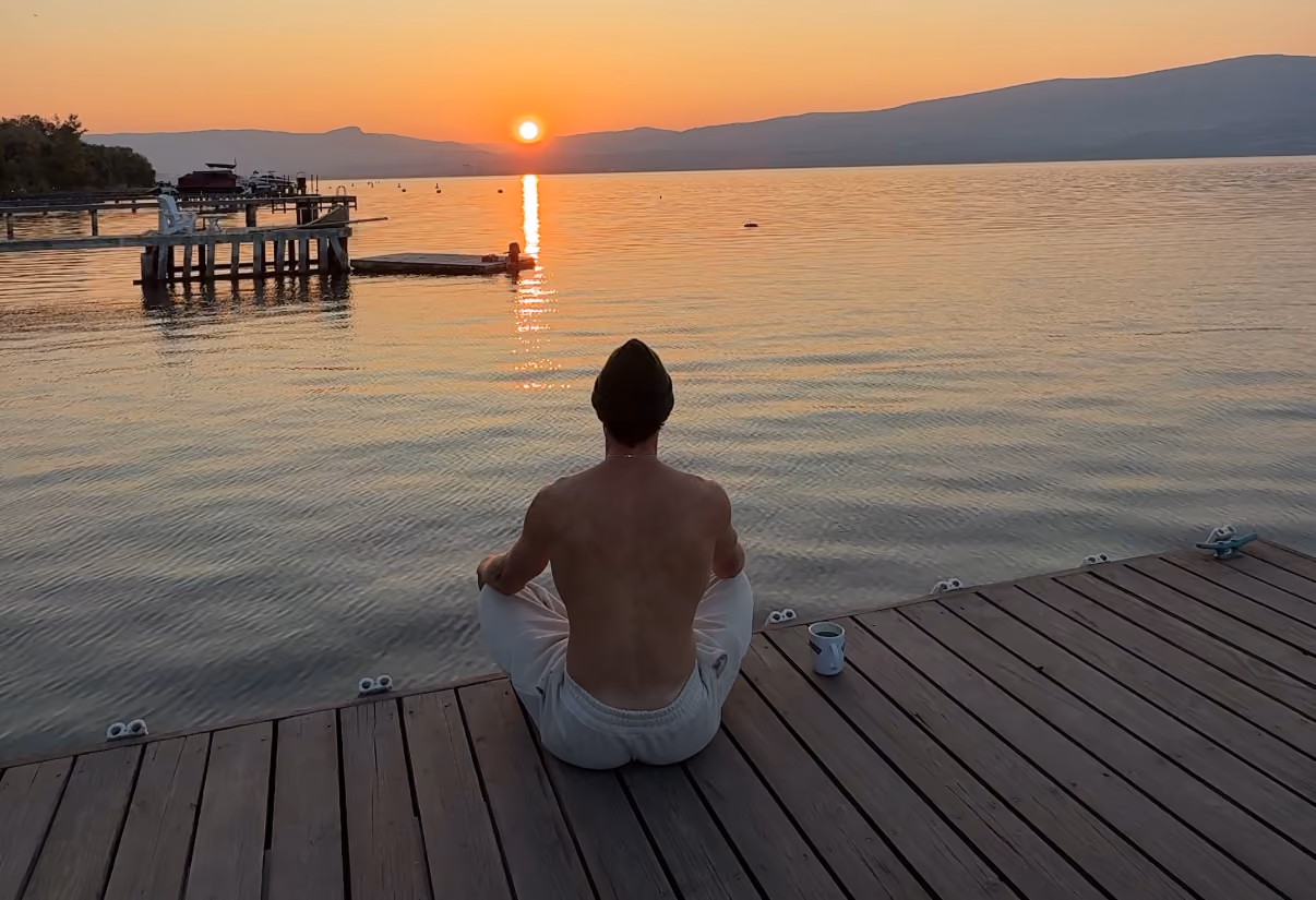 Shirtless man wearing a beanie sitting cross-legged on a dock facing sunset over calm lake waters.