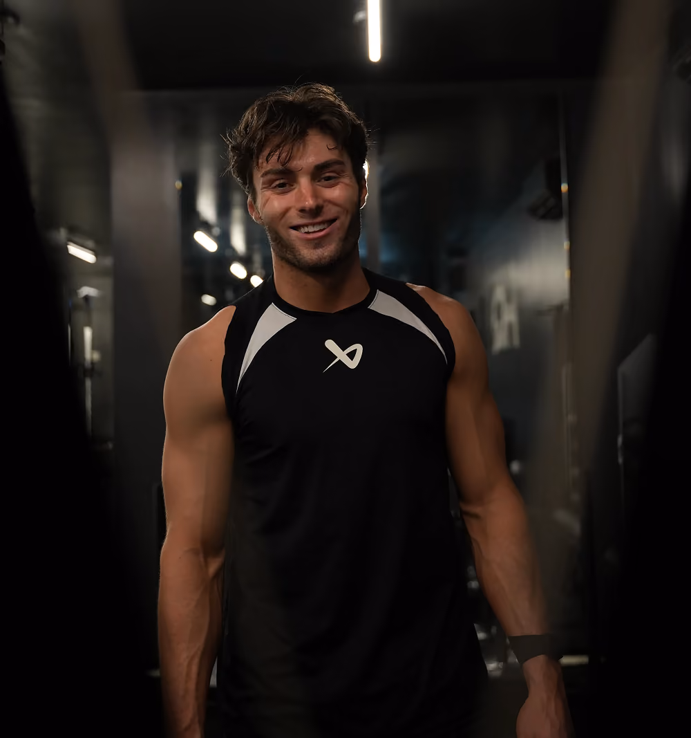 Smiling young man in black sleeveless athletic shirt in a dimly lit gym corridor.