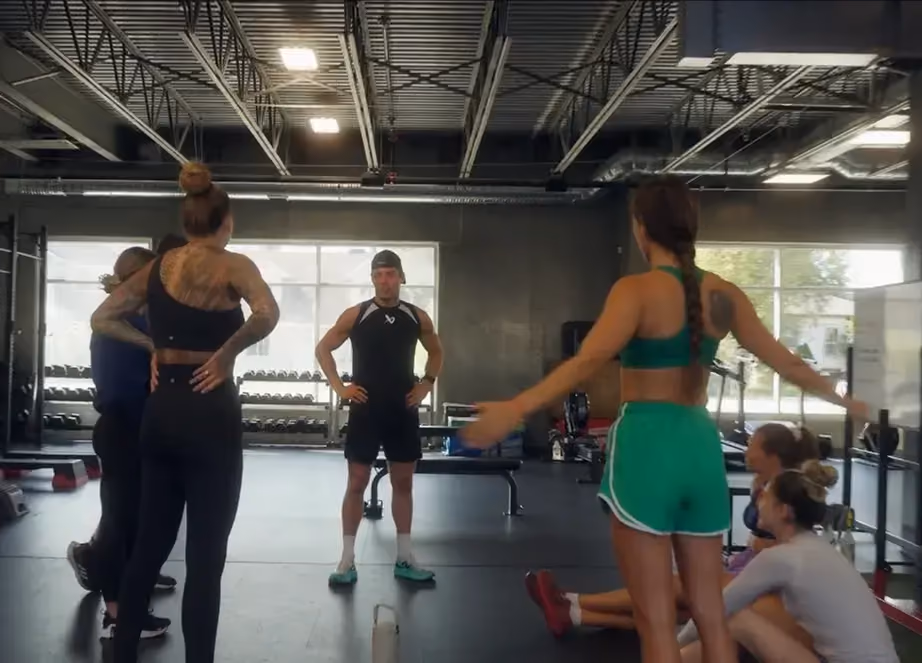 Group of young adults in workout clothes standing and sitting in a gym during a training session.