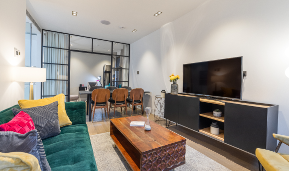 Modern living room with green sofa, wooden table, TV unit, and glass partition to dining area.