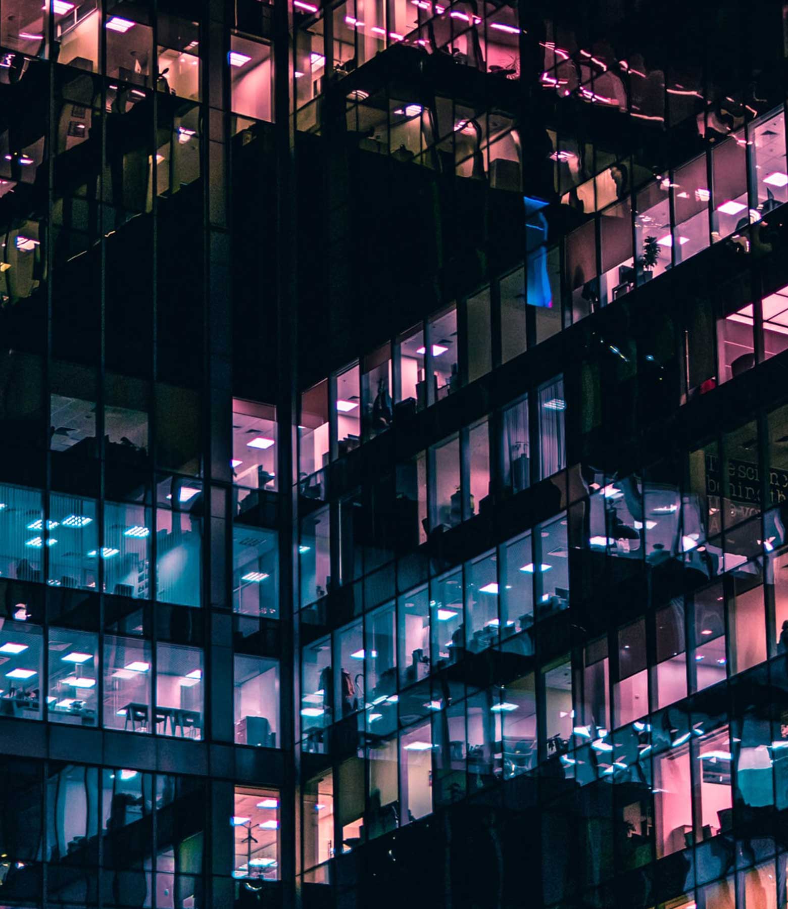 A modern glass building at night, illuminated by vibrant pink and blue interior lights.