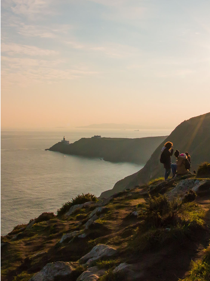 A coastal cliffside at sunset with a lighthouse in the distance and people enjoying the view.