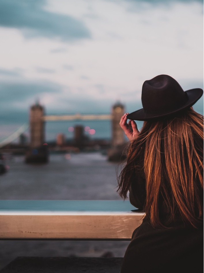 A woman in a black hat gazes at Tower Bridge during dusk, with her long hair illuminated softly.