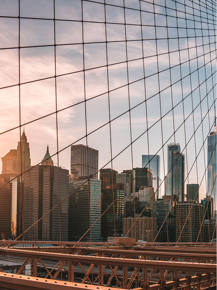 Sunlit city skyline framed by the cables of the Brooklyn Bridge at sunset.