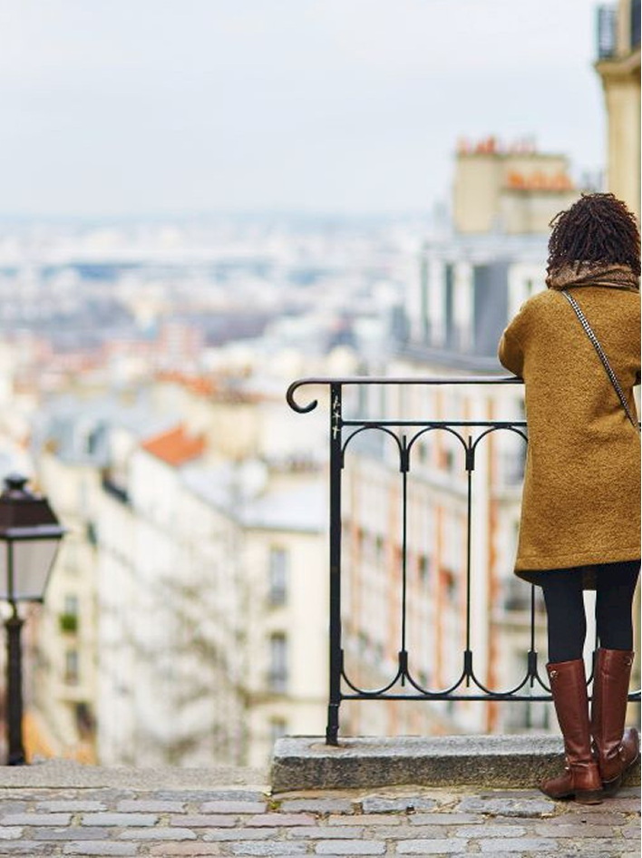 A woman in a brown coat and boots stands at a railing, overlooking a cityscape on a cloudy day.