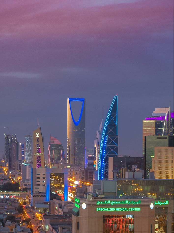 Riyadh skyline at dusk, featuring the Kingdom Centre and Al Anoud Tower illuminated in vibrant lights.
