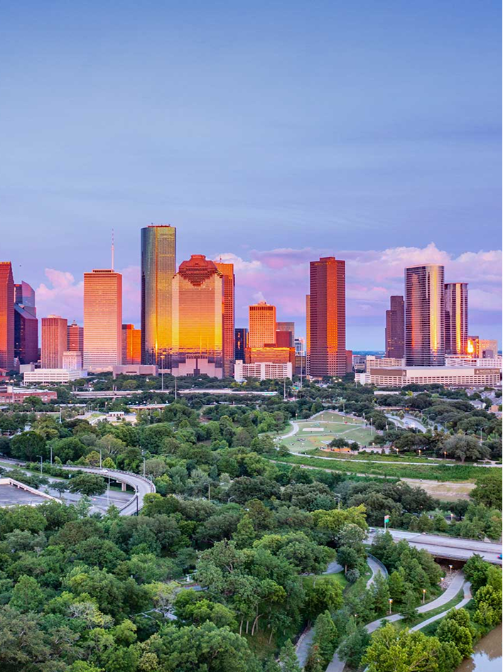 A vibrant city skyline at sunset overlooks lush green parks and winding pathways below.