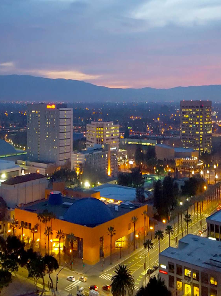 A vibrant cityscape at dusk featuring illuminated buildings, palm-lined streets, and distant hills.
