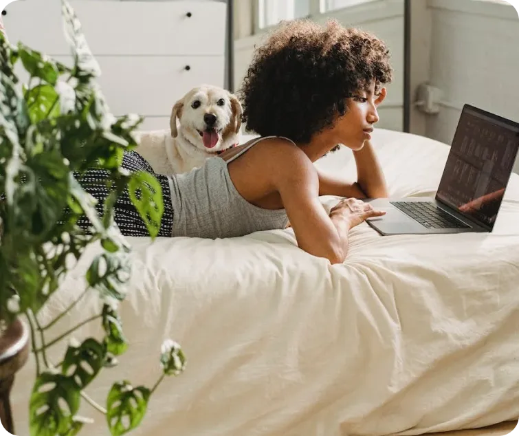 A person lies on a bed using a laptop, accompanied by a happy dog in a bright, cozy room.