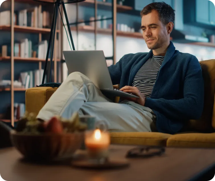 A person sits on a yellow couch, working on a laptop in a cozy room with bookshelves and warm lighting.