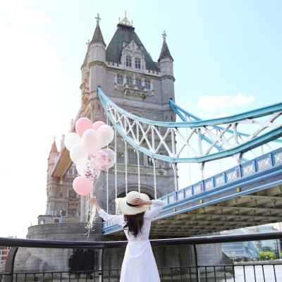A woman in a white dress holds pink and white balloons near London's iconic Tower Bridge.