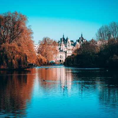 A serene lake reflects autumn trees and a grand historic building under a vibrant blue sky.