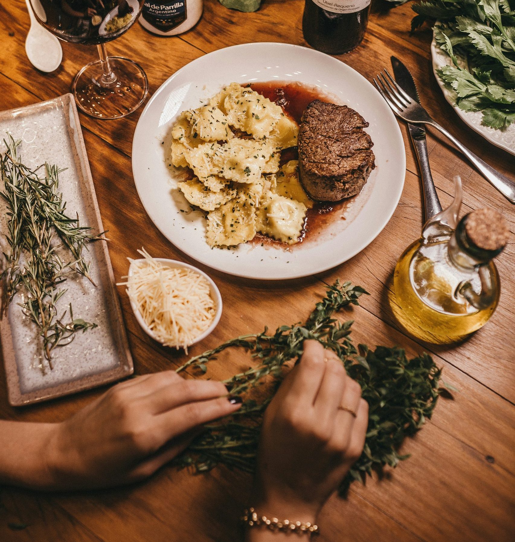 A plated steak with ravioli, fresh herbs, olive oil, and wine on a wooden table setting.