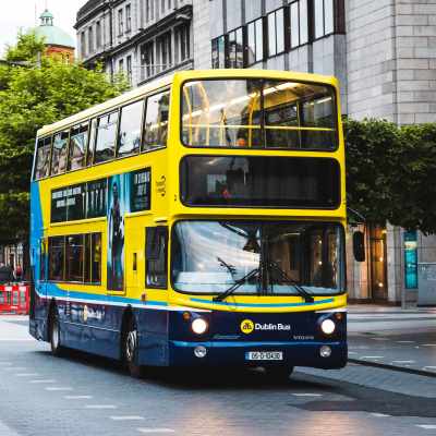 A yellow and blue Dublin Bus drives through a city street lined with trees and buildings.