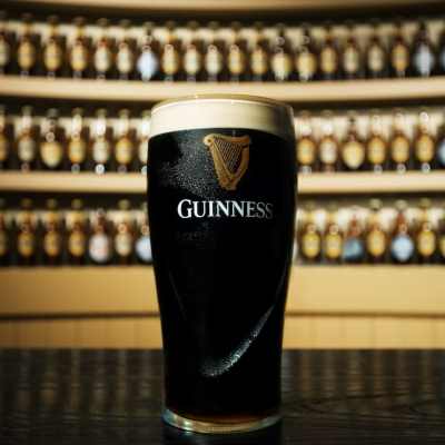 A pint of Guinness stout with a creamy head, set against a backdrop of bottles on shelves.