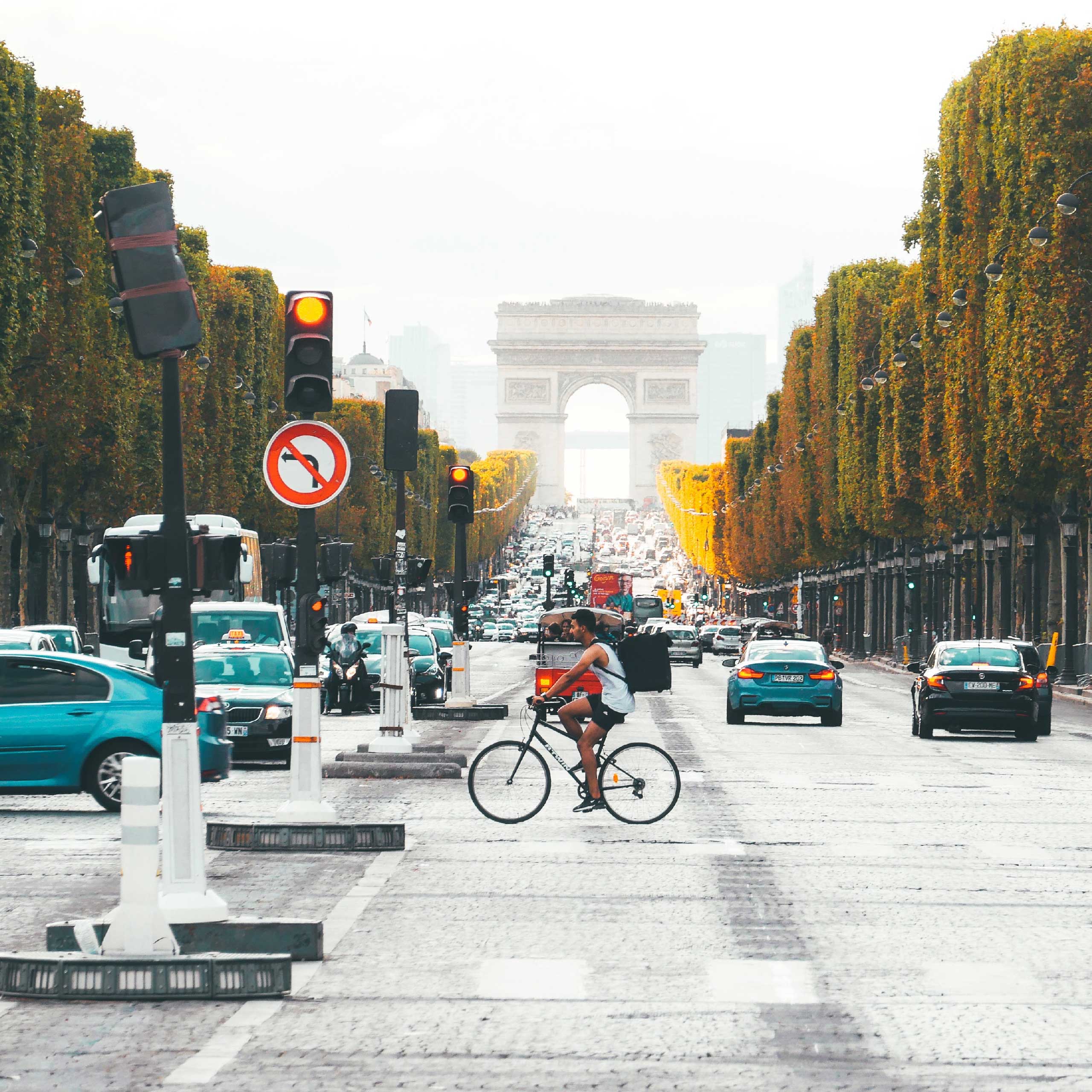 A cyclist crosses a busy street in Paris, with the Arc de Triomphe framed in the background.