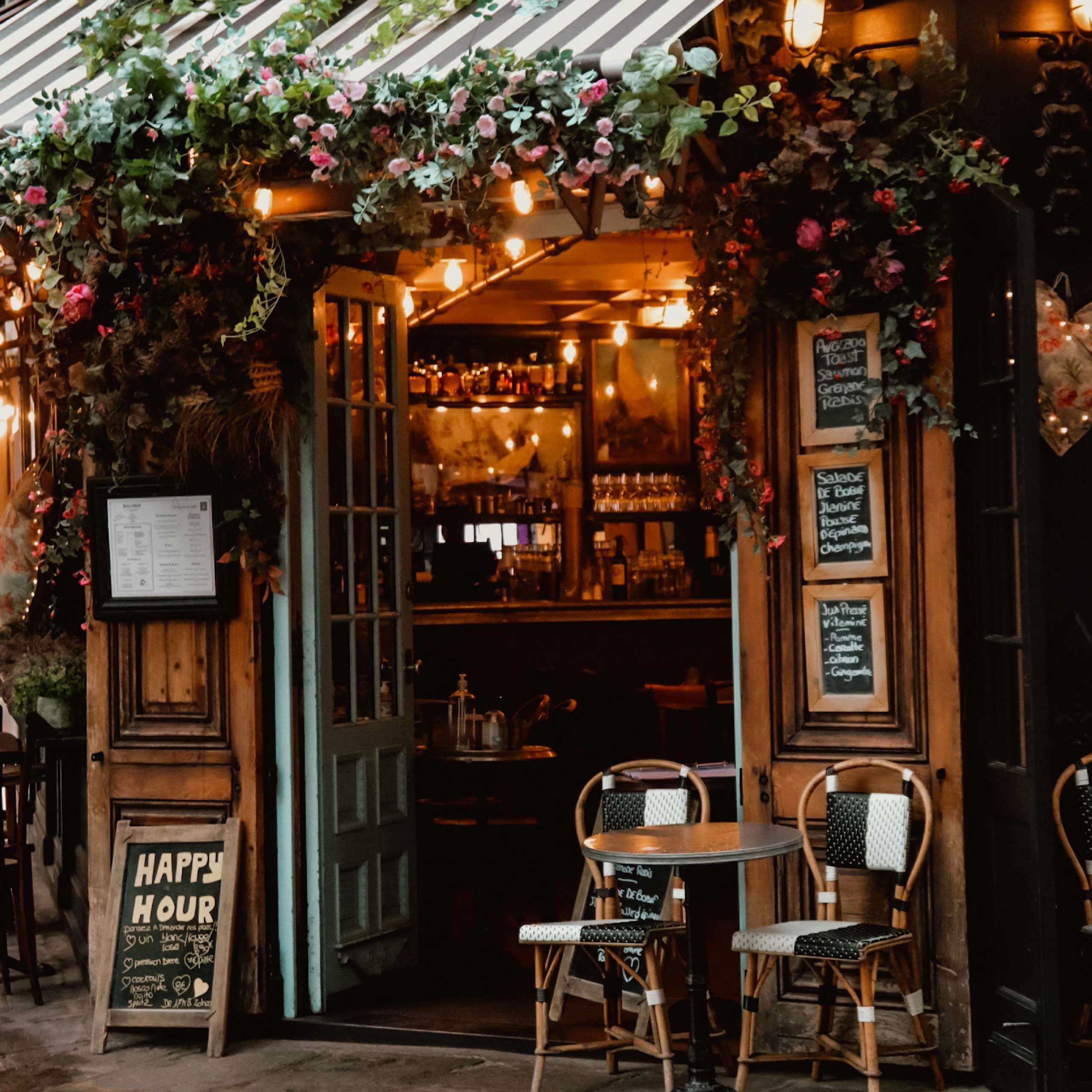 A charming café entrance adorned with flowers, warm lighting, and outdoor seating under a striped awning.
