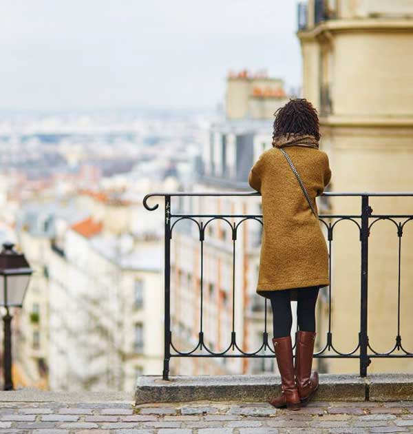 A woman in a brown coat overlooks a cityscape from a wrought-iron railing on a cloudy day.