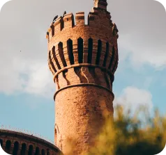 A medieval-style stone tower with crenellations, set against a partly cloudy sky and greenery.
