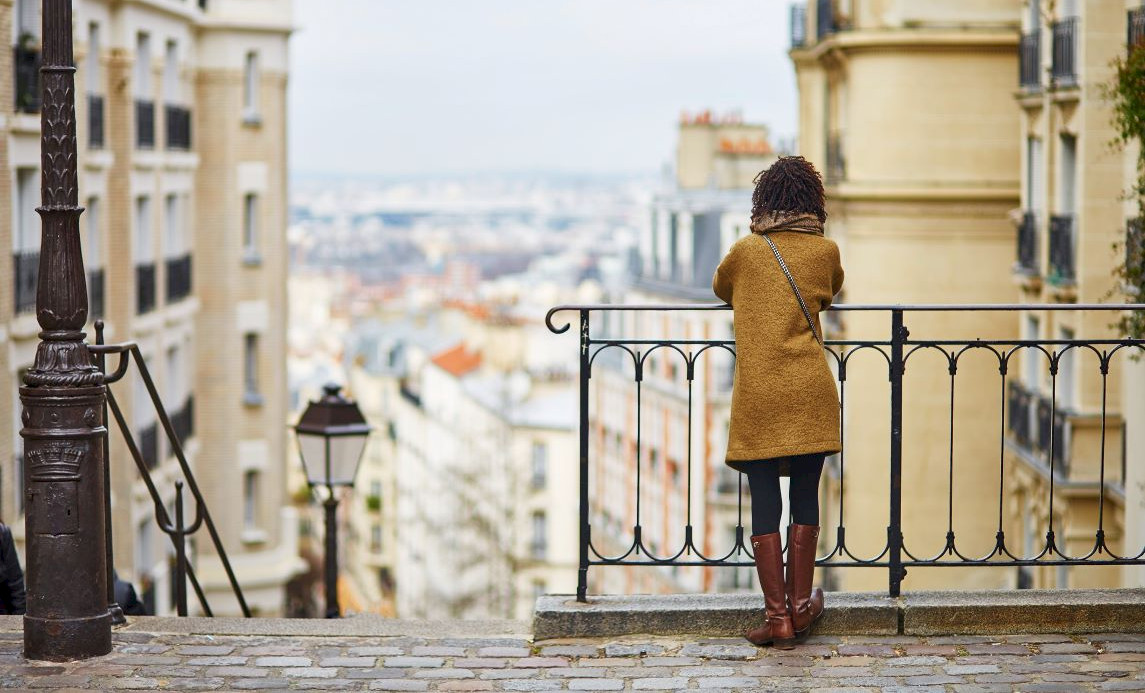 A woman in a brown coat overlooks a cityscape from a railing on a cobblestone street.