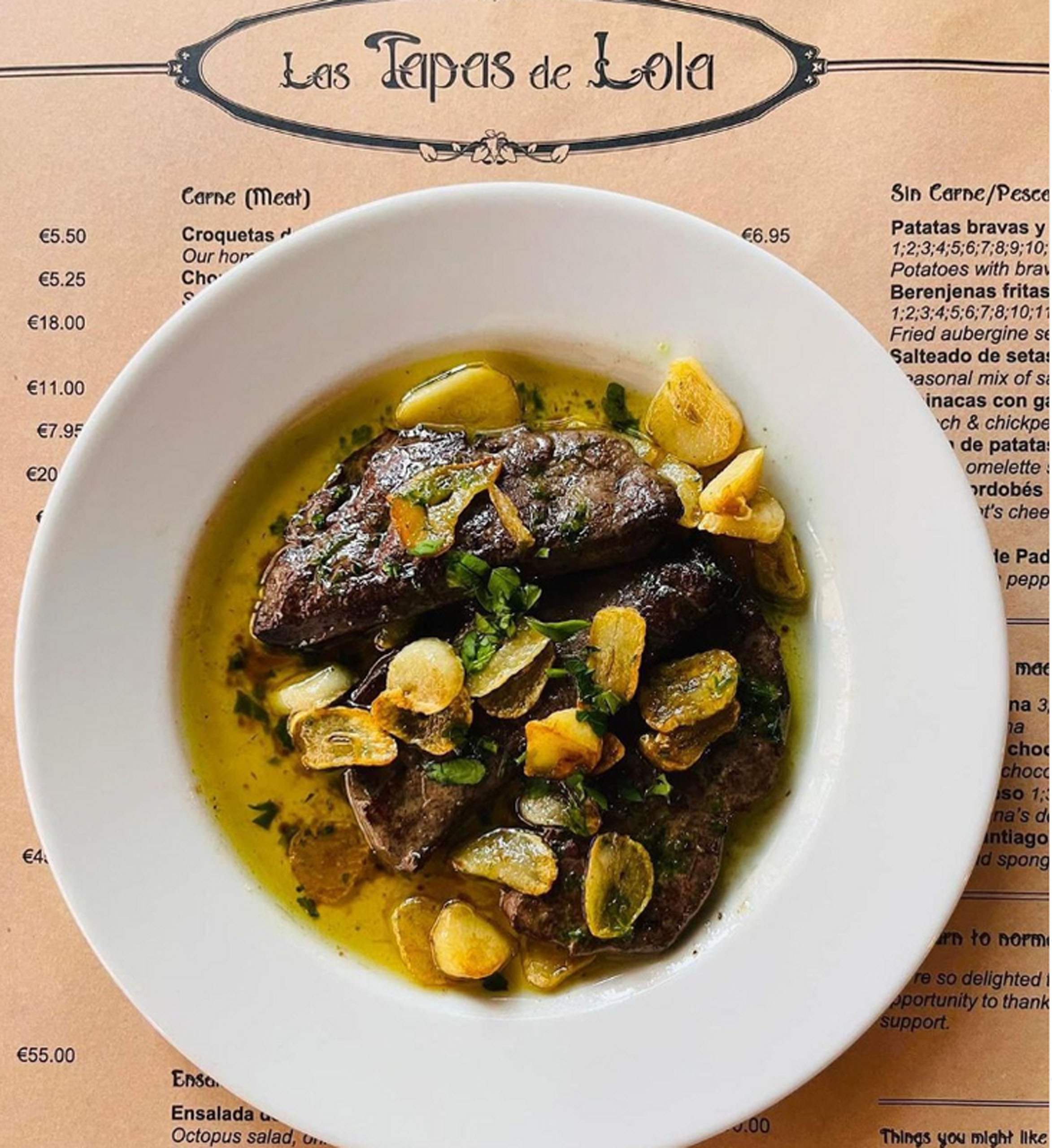 A plate of meat garnished with garlic and herbs, served on a menu from Las Tapas de Lola restaurant.