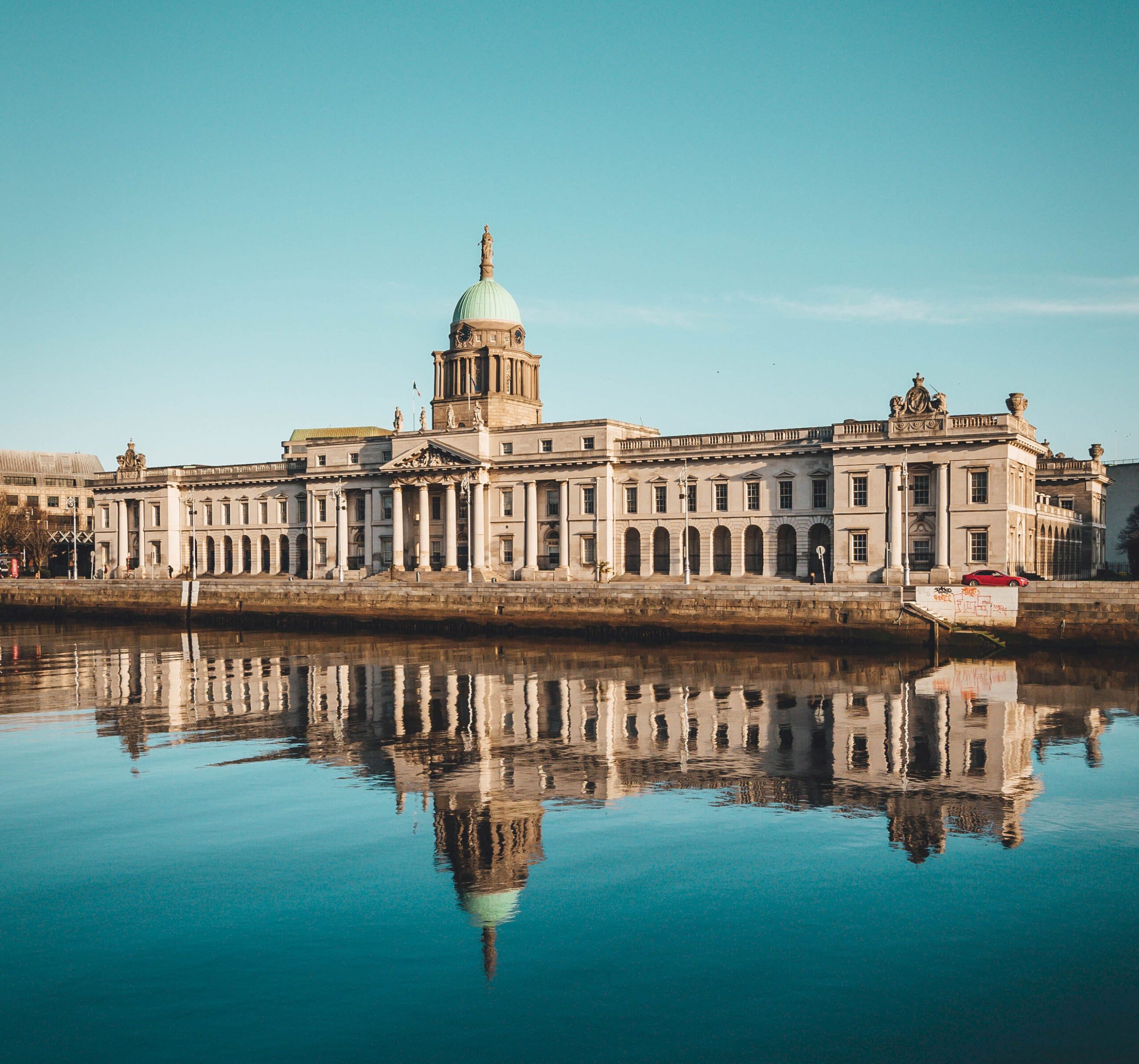 The Custom House in Dublin, Ireland, reflects on the calm waters under a clear blue sky.
