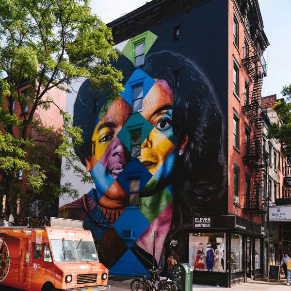 A vibrant mural covers a building facade, surrounded by trees, shops, and an orange food truck.