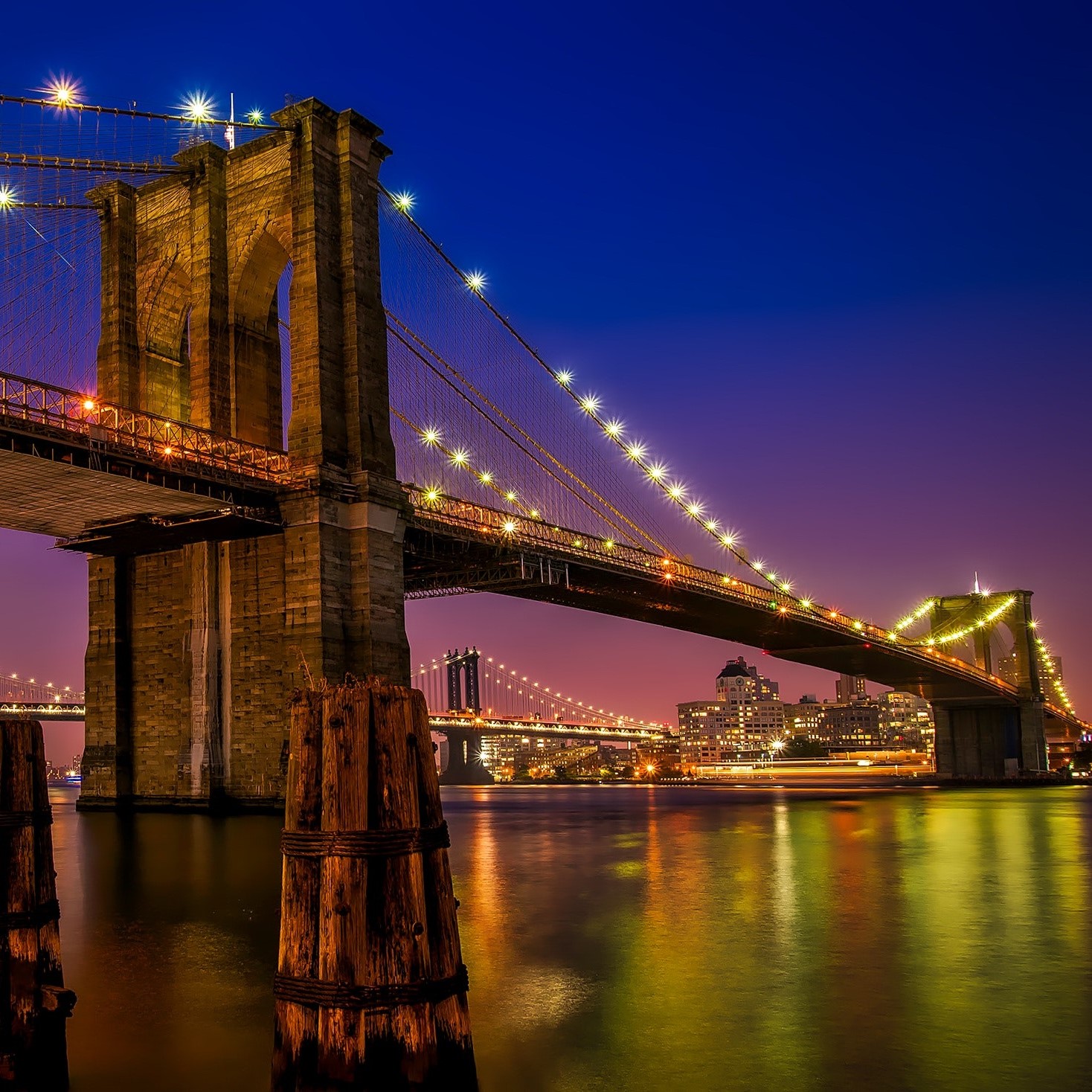 The Brooklyn Bridge illuminated at night, spanning the East River with a vibrant cityscape backdrop.