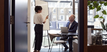 A woman presents at a whiteboard to a seated man in a modern office with large windows.